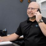 Homme chauve portant des lunettes et un polo noir parlant au téléphone tout en écrivant dans un carnet au bureau