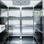 Étagères métalliques vides dans une chambre froide industrielle avec quelques contenants plastiques et produits alimentaires