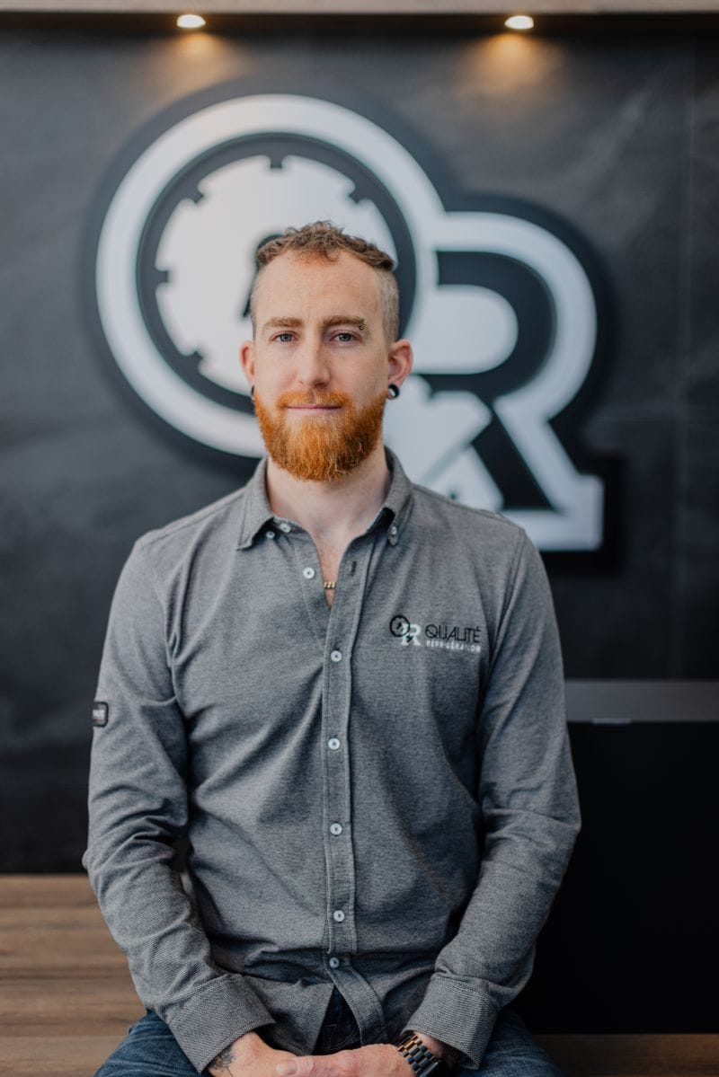 Homme avec barbe rousse et chemise grise assis devant un mur noir avec un logo d'entreprise