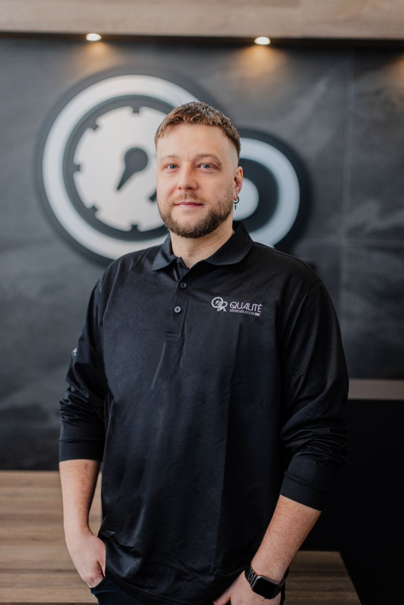 Homme portant un polo noir avec logo Qualité Réfrigération Inc., posant devant un mur gris avec un grand logo circulaire blanc et noir.