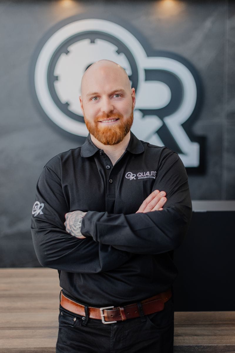 Homme souriant avec barbe rousse et tête chauve portant un polo noir avec le logo Qualité Réfrigération devant un mur gris avec un grand logo en arrière-plan