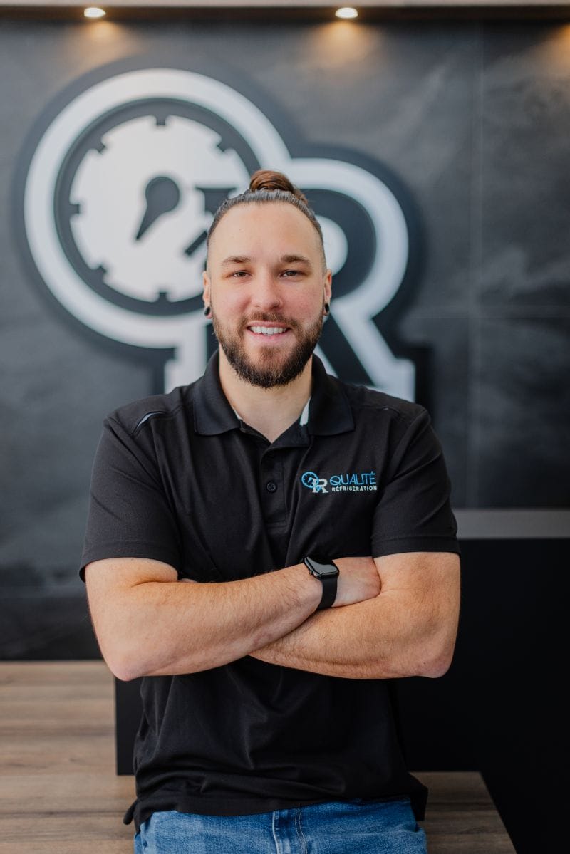 Homme souriant portant un polo noir avec le logo Qualité Réfrigération devant un mur avec un grand logo en arrière-plan