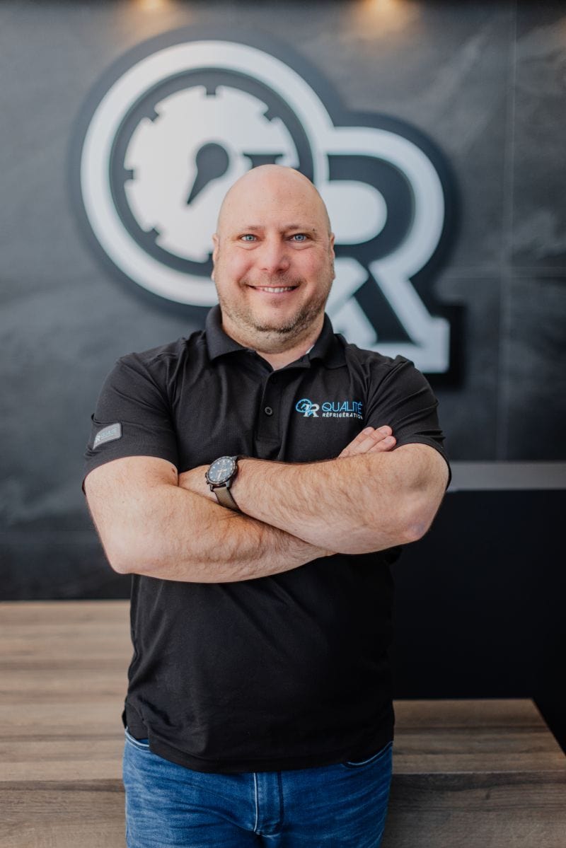 Homme souriant avec les bras croisés portant un polo noir avec logo de Qualité Réfrigération devant un arrière-plan de bureau moderne