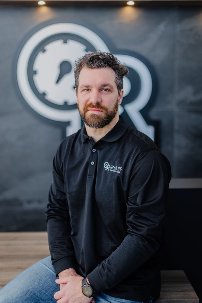 Homme avec barbe portant un polo noir avec logo assis devant un mur avec un grand logo flou