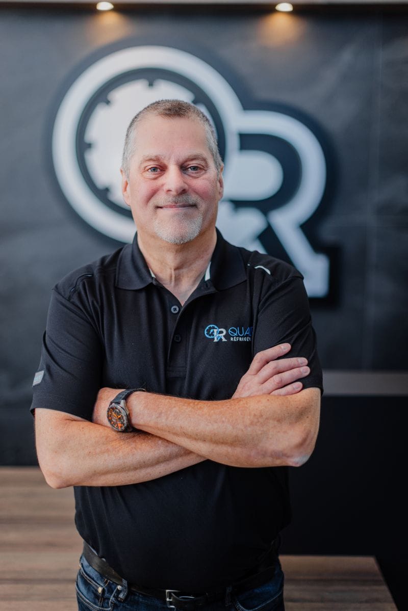 Homme souriant avec les bras croisés portant un polo noir de marque Qua Réfrigération devant un logo d'entreprise flou