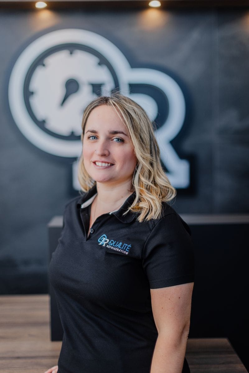 Portrait d'une femme souriante portant un polo noir avec le logo Qualité Réfrigération, devant un mur sombre avec une horloge floue en arrière-plan.