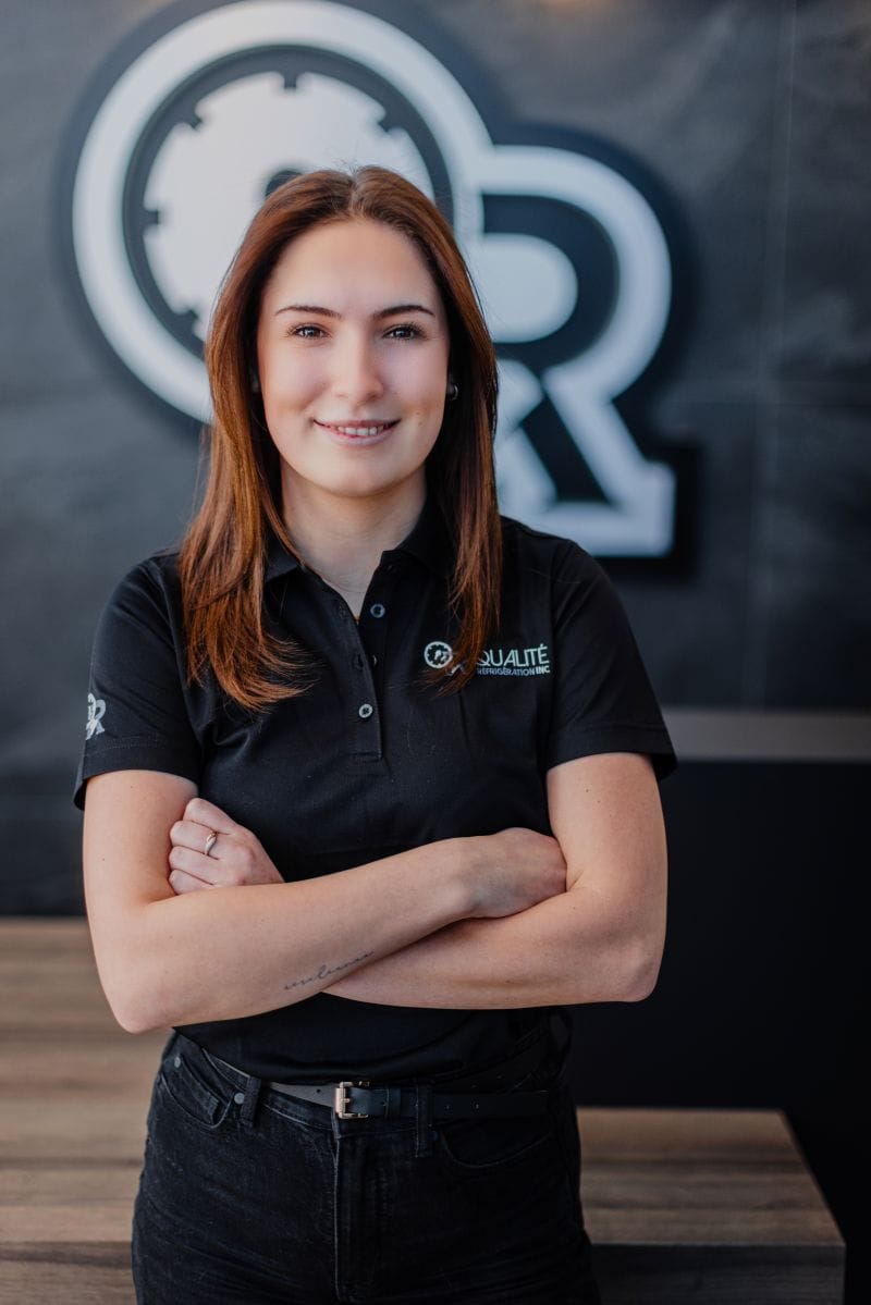 Jeune femme souriante en polo noir avec logo d'entreprise, bras croisés devant un mur sombre avec un grand logo blanc