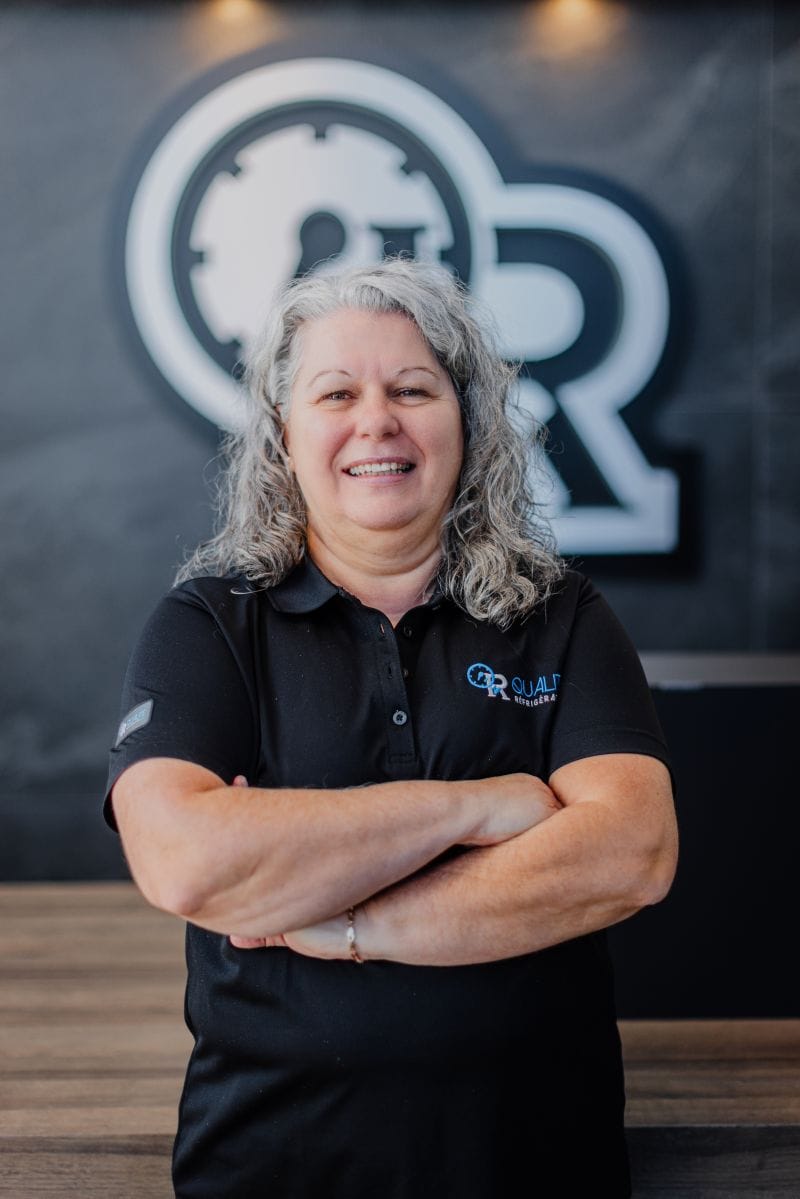 Femme souriante aux cheveux gris portant un polo noir avec logo Q&R Qualité Réfrigération, debout devant un mur avec grand logo flou en arrière-plan