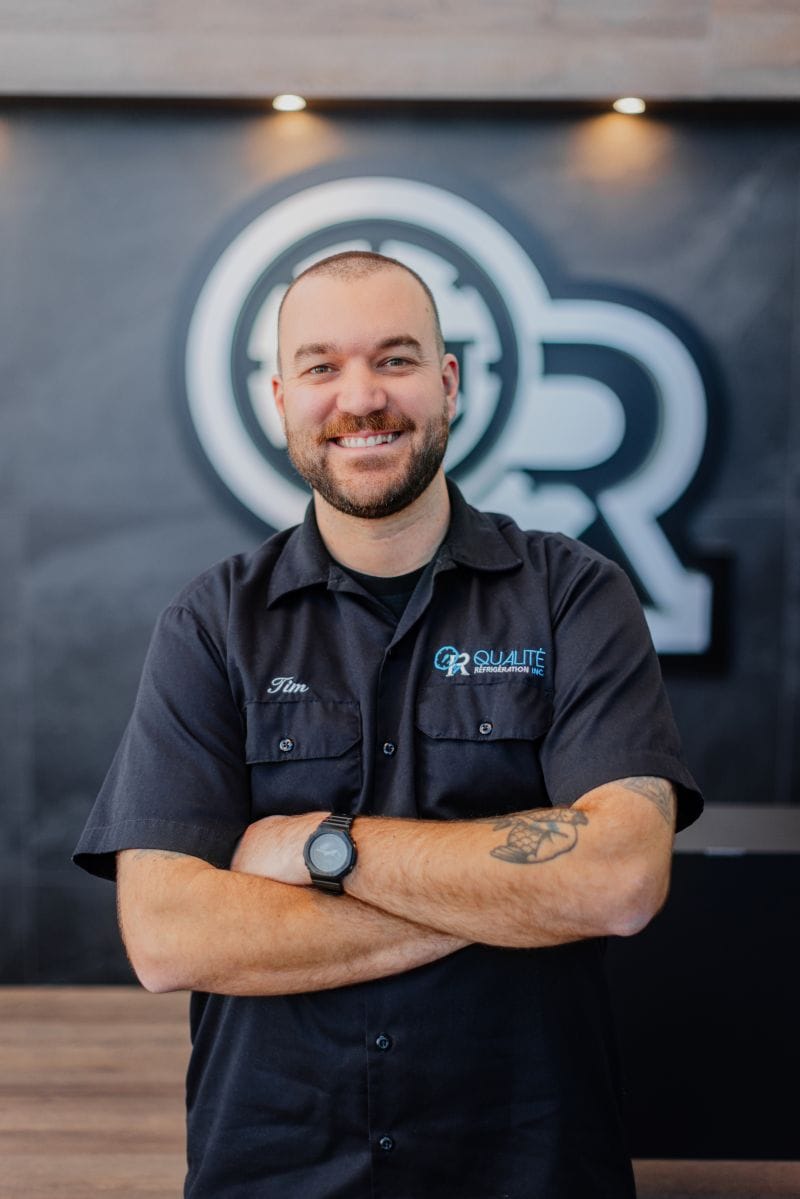 Homme souriant portant une chemise noire brodée avec le logo de Qualité Réfrigération inc. et un tatouage visible sur le bras